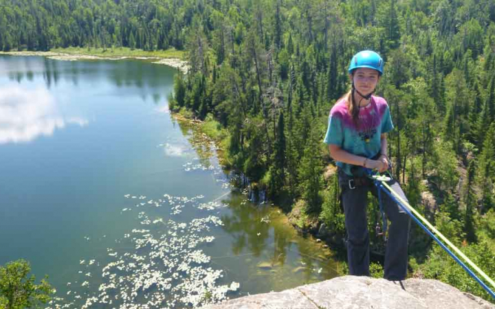 A young person wearing safety gear is secured by ropes as they stand on the edge of a cliff, high above a body of water and a forrest. 